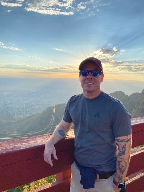 Man standing with hand on bridge with mountains behind him with sunlight.