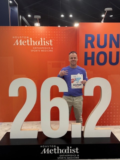 Man standing behind a 26.2 sign holding running bib. Hosted by Houston Methodist.